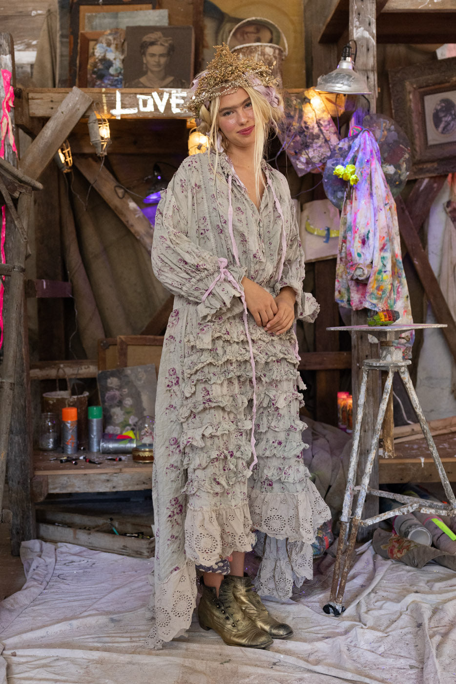 Woman in a floral dress standing in a rustic room with wooden walls and various items.