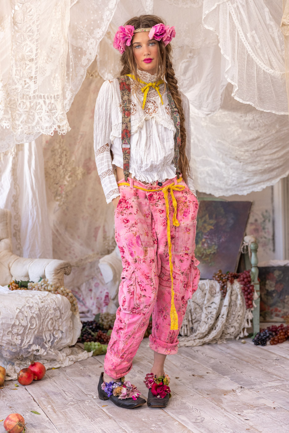 Woman in Magnolia Pearl artisan clothing, whimsical floral pants, vintage lace blouse, floral crown, and artistic shoes, standing in romantic vintage room.