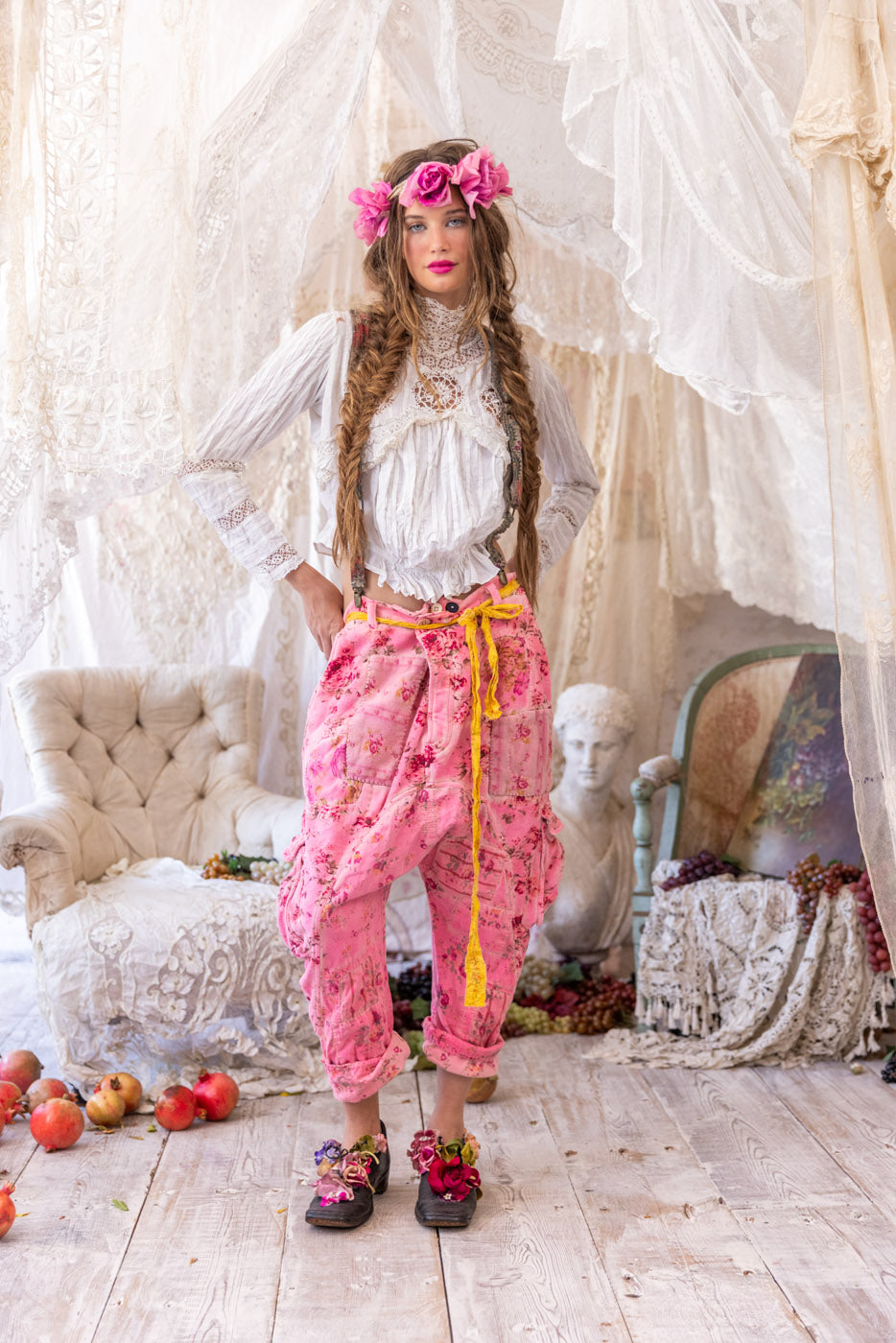 Woman in Magnolia Pearl artisan clothing, whimsical floral pants, vintage lace blouse, floral crown, and artistic shoes, standing in romantic vintage room.