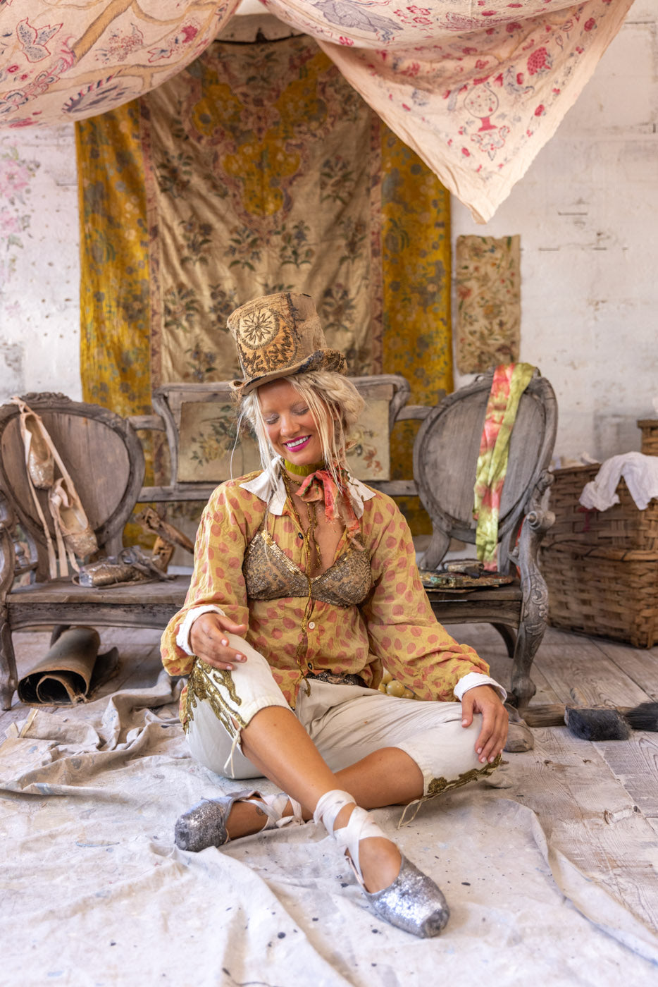 Woman in whimsical Magnolia Pearl artisan clothing, vintage top hat, and ballet shoes, seated on antique bench amid eclectic boho decor