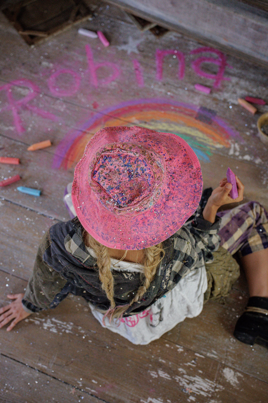 Woman in vintage Magnolia Pearl artisan clothing, pink whimsical hat, drawing rainbow with chalk on rustic wood floor