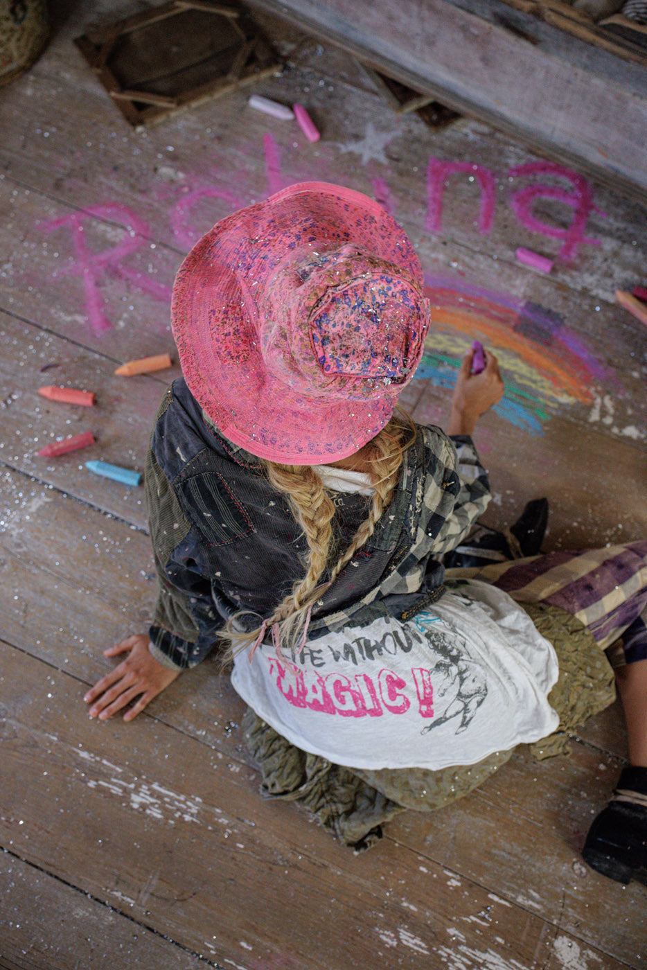 Woman in Magnolia Pearl artisan vintage clothing, pink hat, drawing rainbow with chalk on rustic wood floor