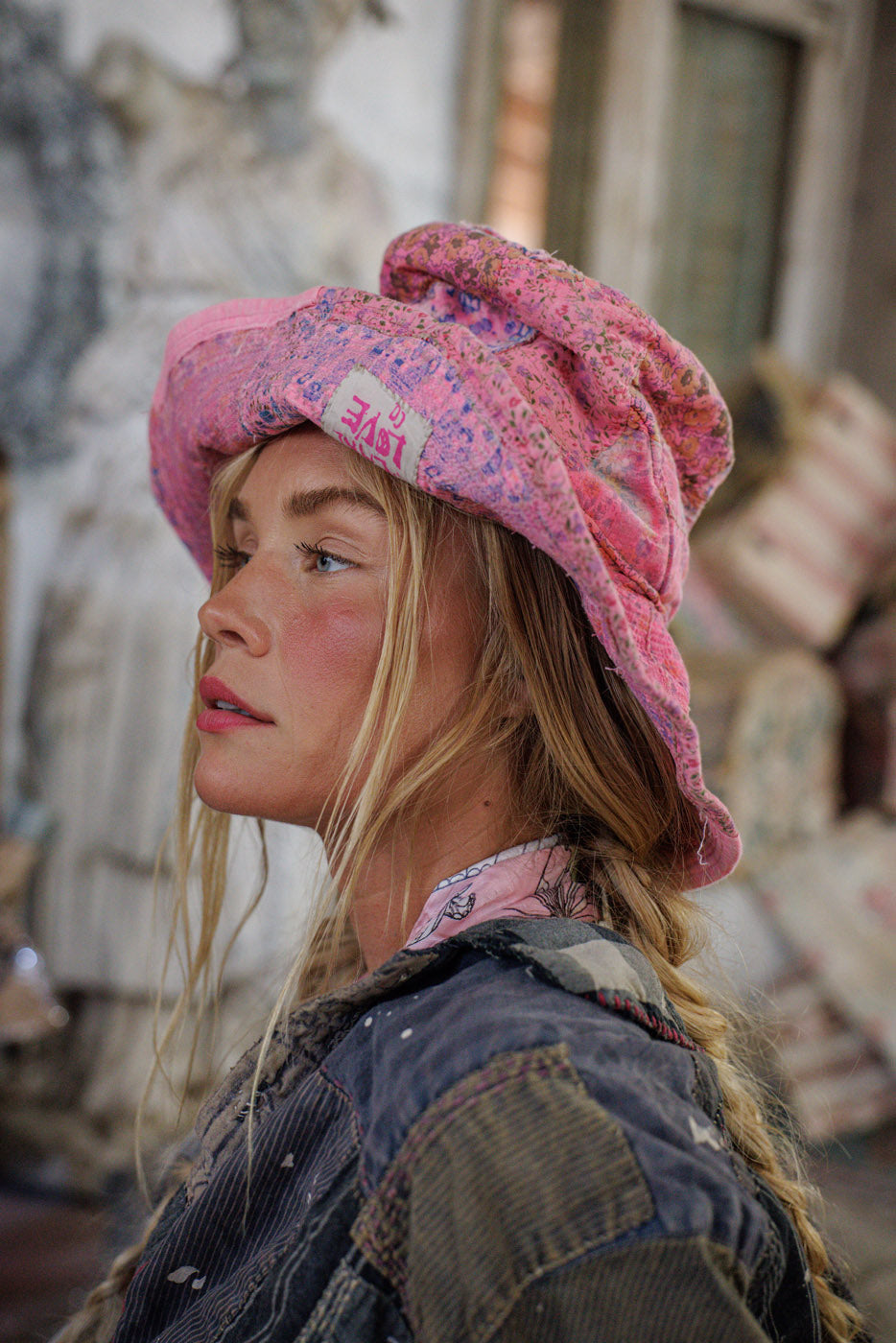 Woman in Magnolia Pearl artisan pink floral hat and vintage patchwork jacket, whimsical style.
