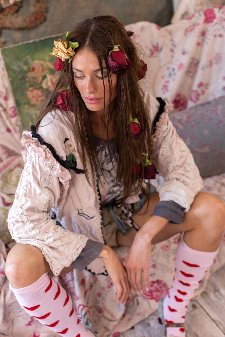 Woman in Magnolia Pearl vintage artisan jacket with whimsical rose accents, sitting on floral fabric.