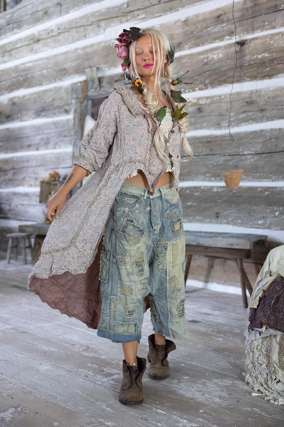 Woman in Magnolia Pearl artisan vintage-style jacket and distressed jeans with whimsical floral hair, rustic setting