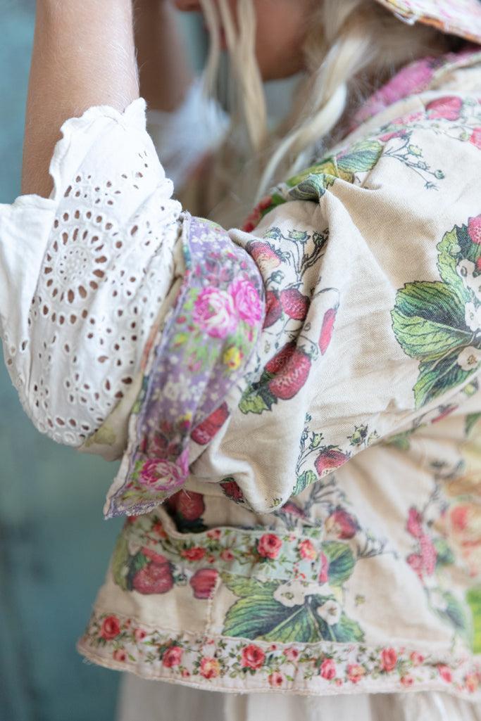 Woman in Magnolia Pearl whimsical vintage artisan clothing, floral embroidered jacket, lace dress, colorful hat, seated on rustic wood floor