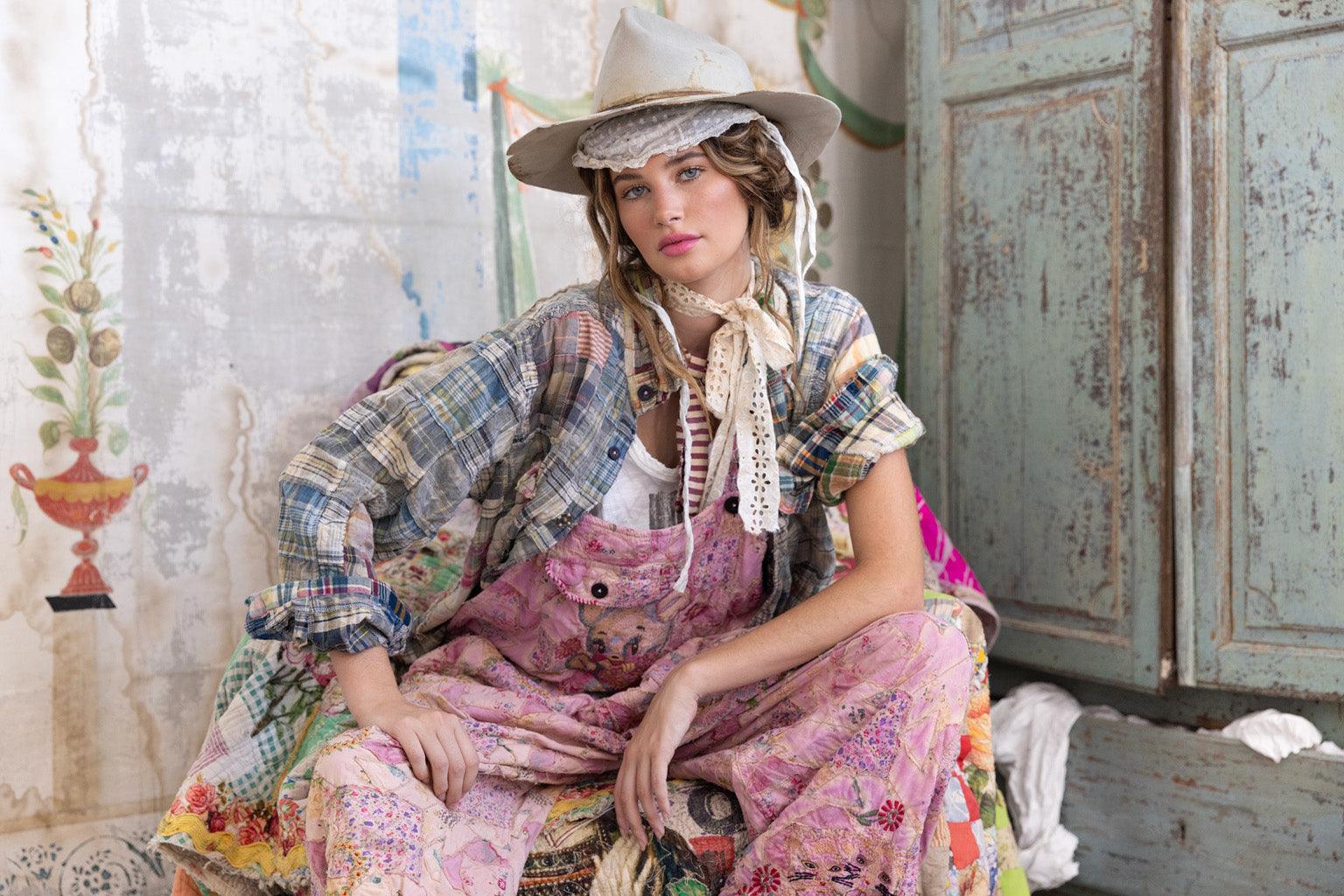 Woman in Magnolia Pearl pink patchwork overalls, plaid shirt, vintage hat, standing by antique armoire and colorful patchwork sofa in whimsical artisan setting