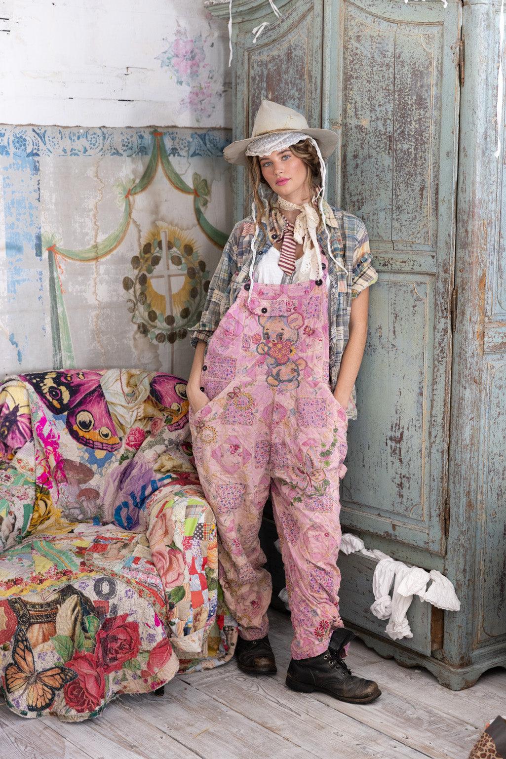 Woman in Magnolia Pearl pink patchwork overalls, plaid shirt, vintage hat, standing by antique armoire and colorful patchwork sofa in whimsical artisan setting