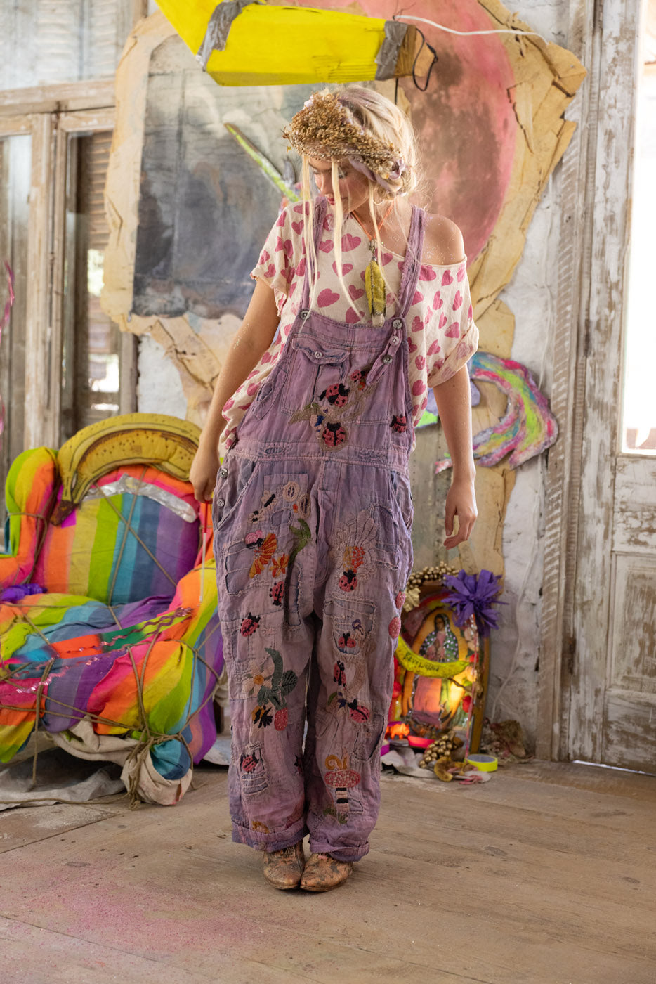 Person wearing a t-shirt with hearts and purple ladybug floral overalls in a colorful, artistic setting.