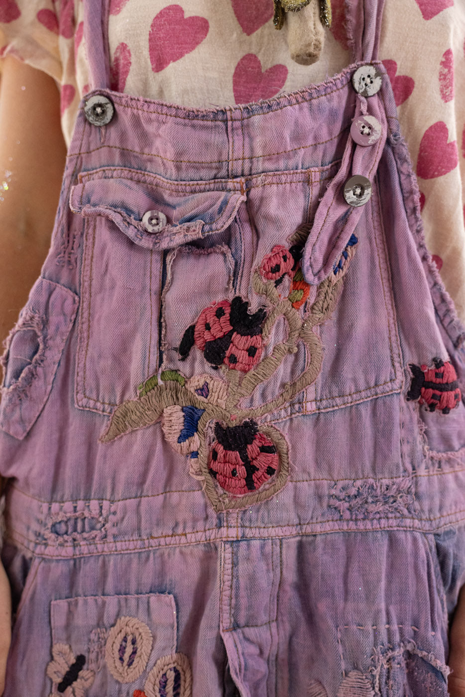 Purple denim overalls with embroidered ladybug design on a person wearing a patterned heart shirt.