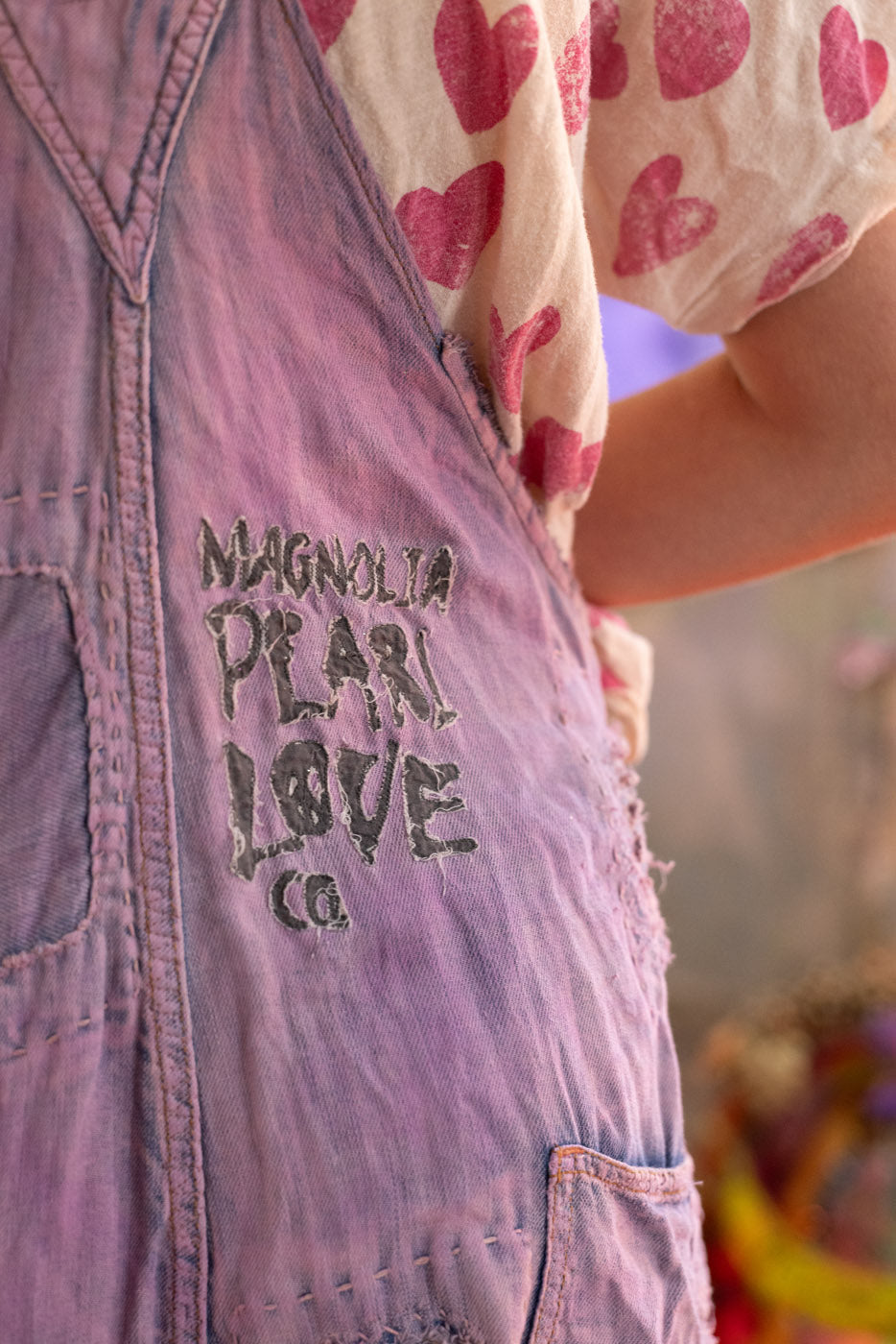 Purple denim overalls with 'Magnolia Pearl Love Co.' branding, worn over a heart-patterned shirt.