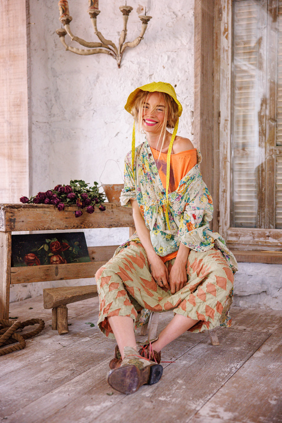 Woman in Magnolia Pearl artisan vintage floral top and patchwork pants, yellow hat, rustic room