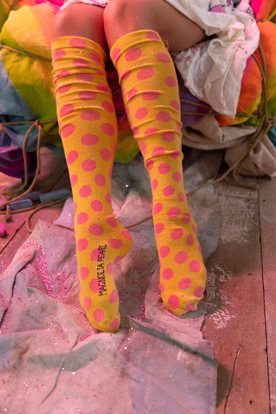 Yellow knee-high socks with pink polka dots worn by a person sitting on colorful fabric.