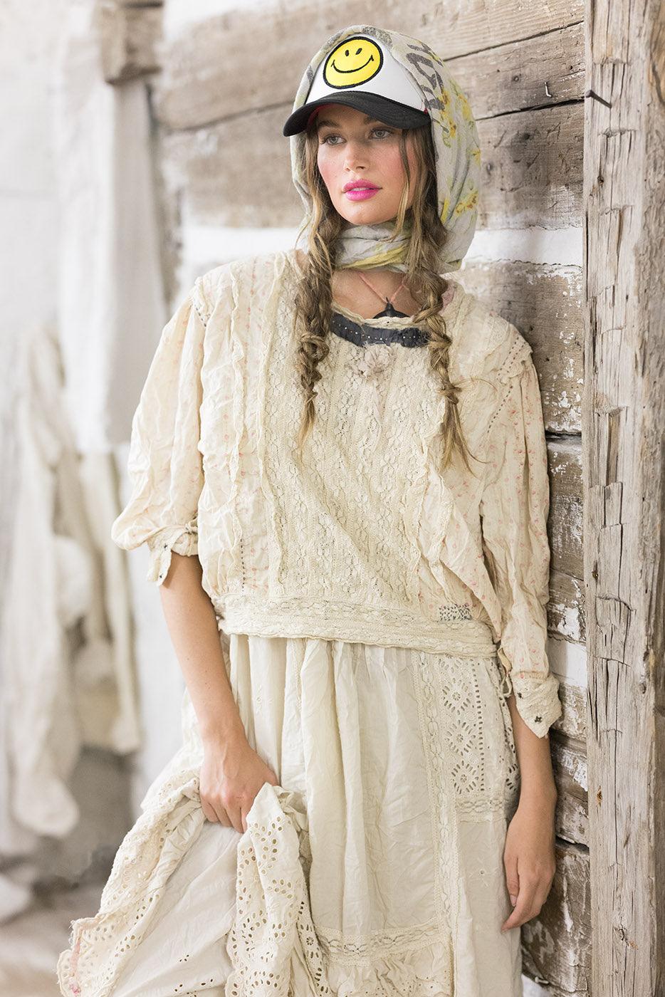 Woman in Magnolia Pearl artisan vintage lace dress, whimsical smiley cap, rustic wood backdrop