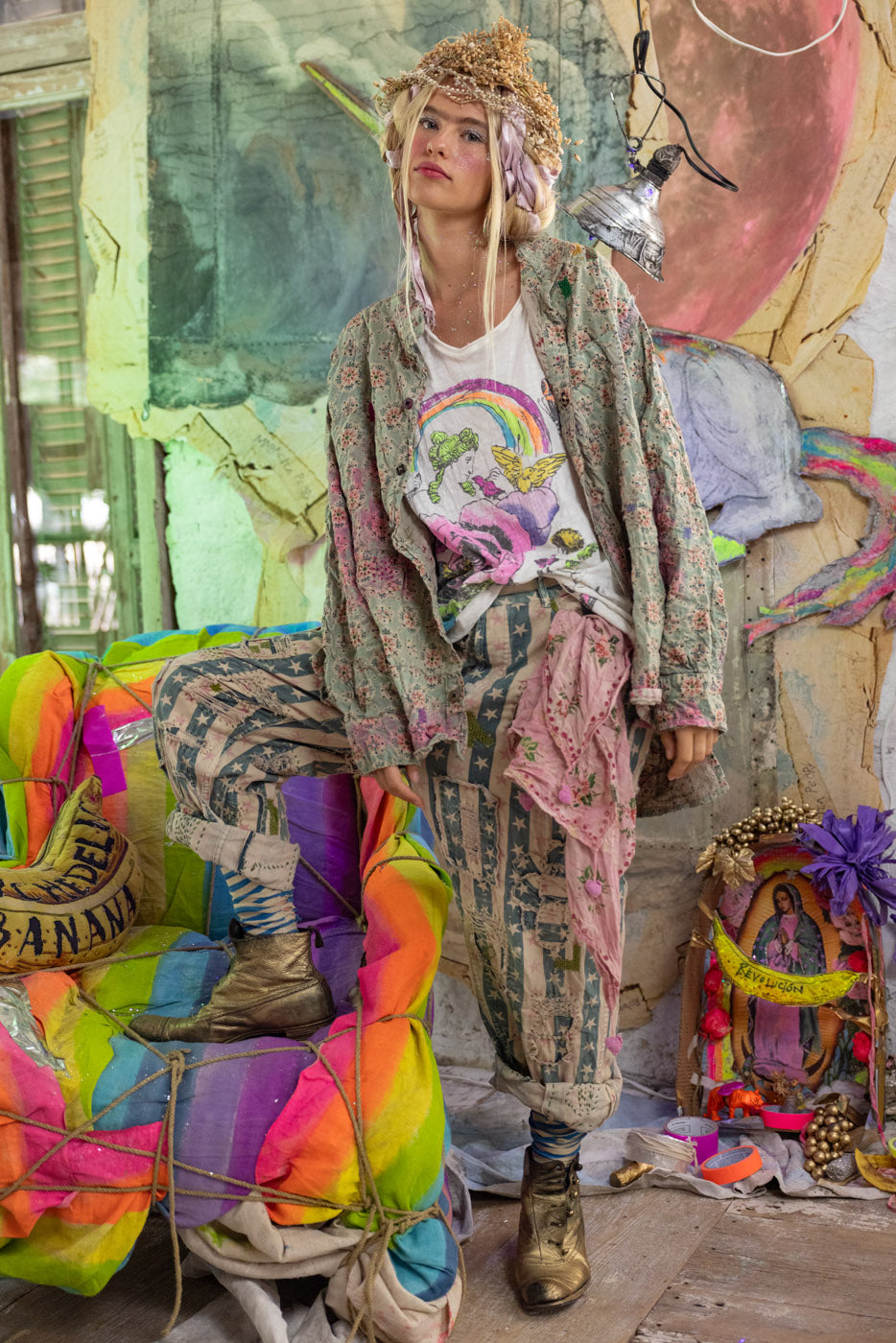 Person in a colorful, eclectic setting with a vibrant mural in the background wearing a rainbow printed t-shirt and star patterned pants and a floral button down shirt.