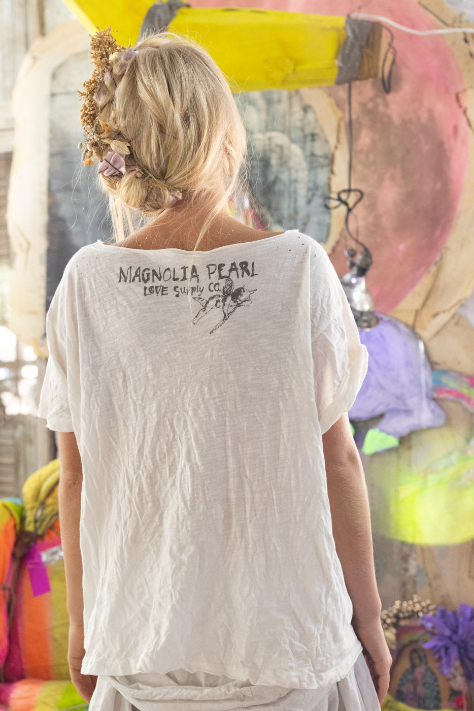Woman wearing a white t-shirt with 'Magnolia Pearl' branding, standing in front of colorful abstract art.