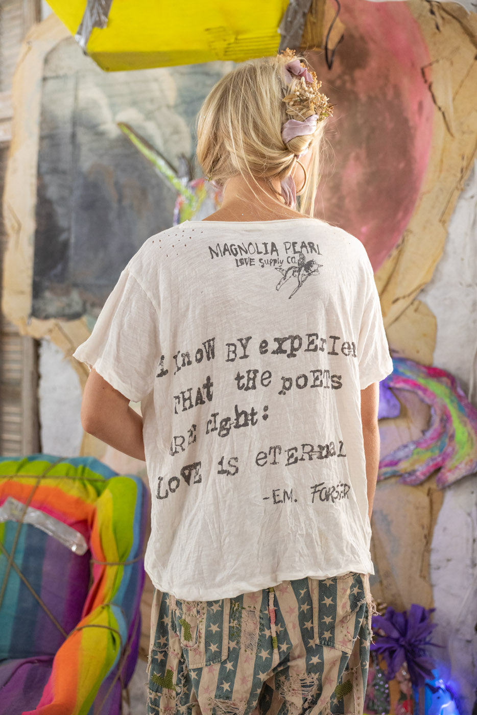 Person wearing a white t-shirt with text, standing in front of a colorful abstract art background.