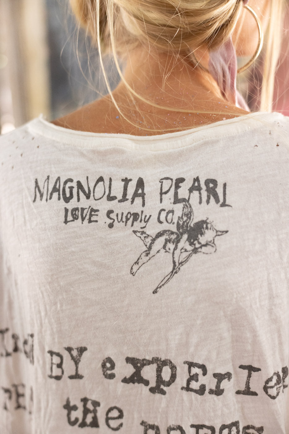 White t-shirt with 'Magnolia Pearl Love Supply Co' text and graphic on a blurred background
