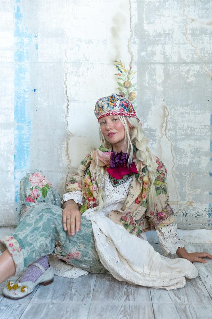 Woman in Magnolia Pearl whimsical vintage artisan clothing, floral embroidered jacket, lace dress, colorful hat, seated on rustic wood floor