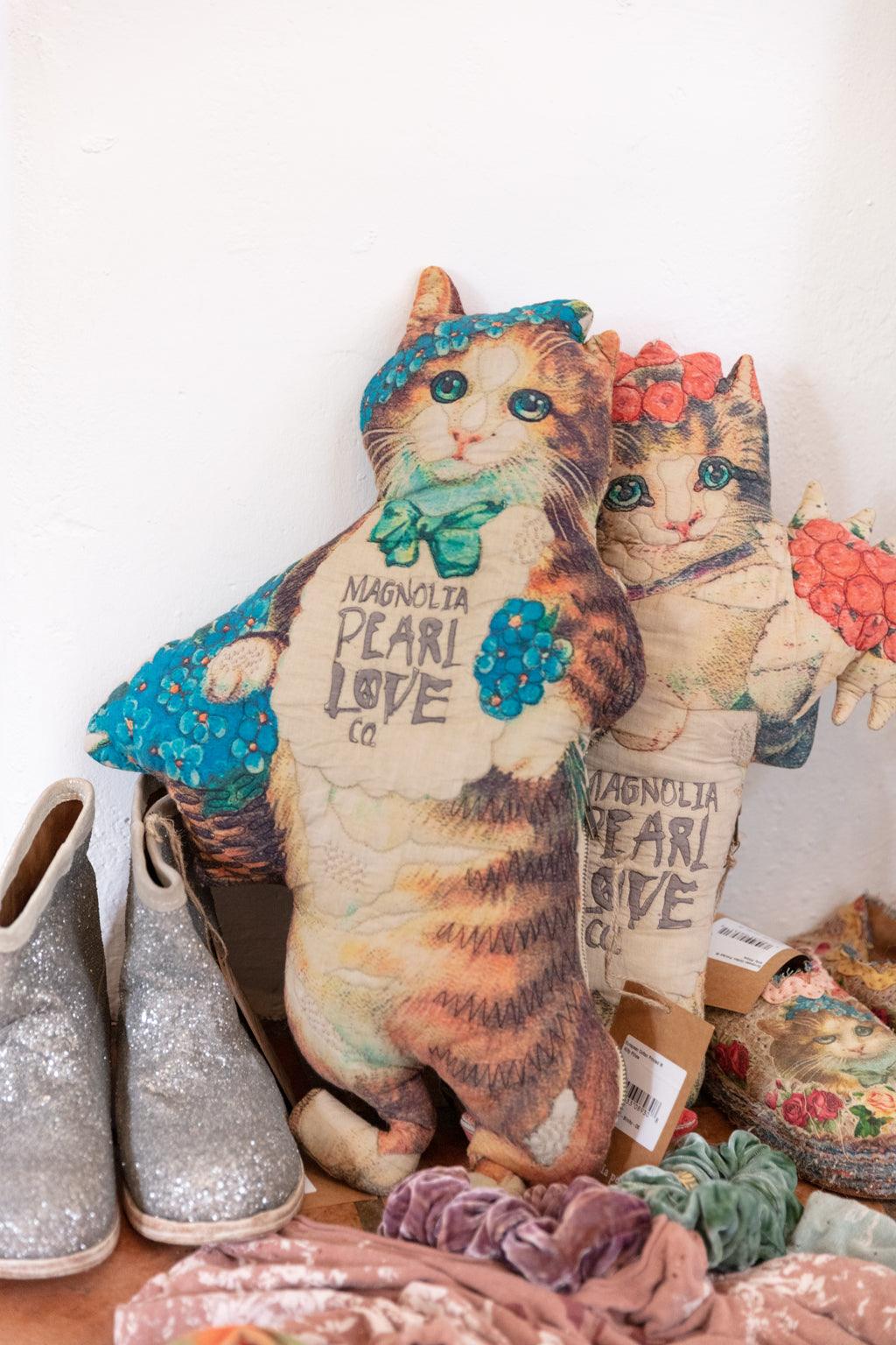 Magnolia Pearl artisan cat pillows with whimsical vintage designs, next to glitter boots and floral accessories.