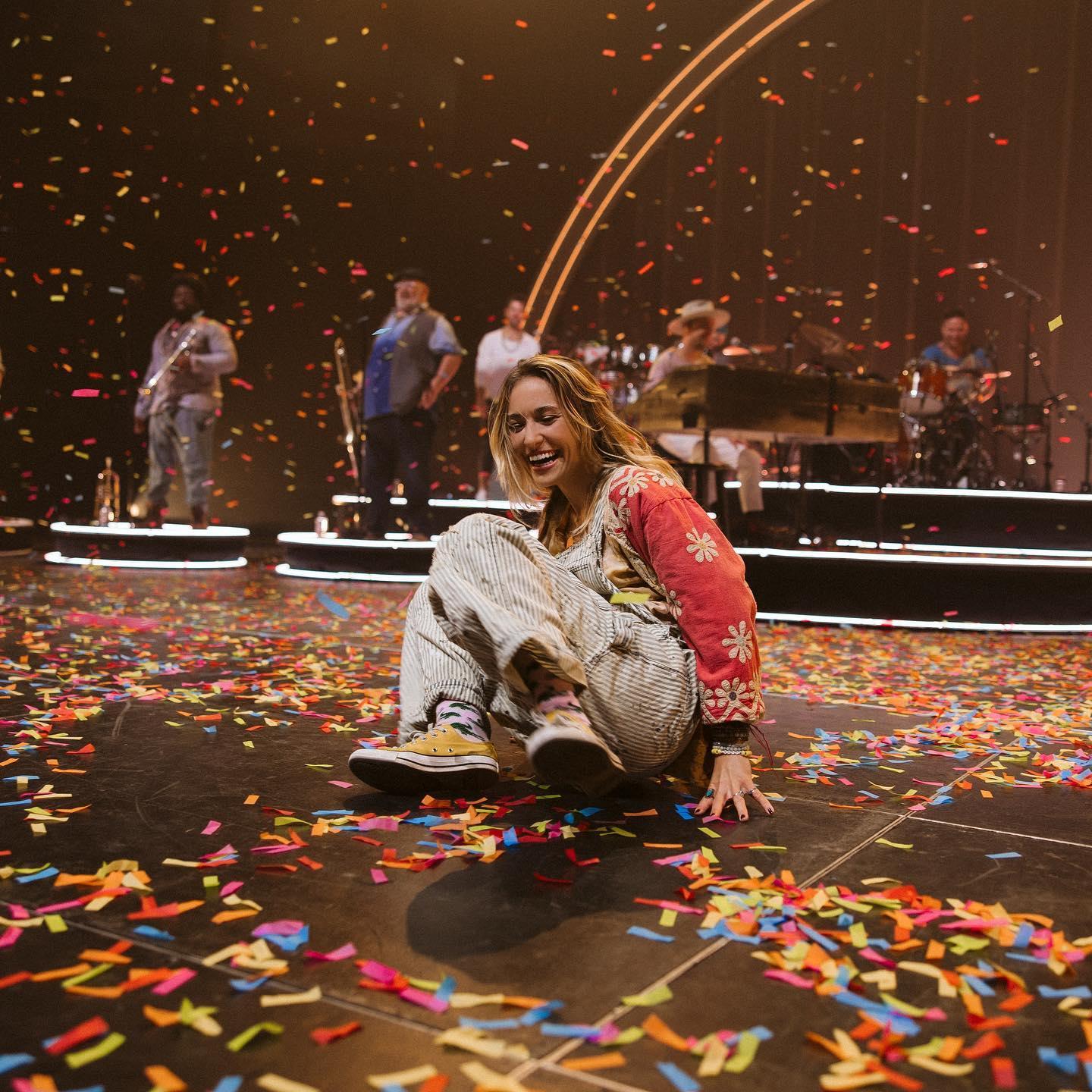 Woman wearing Magnolia Pearl artisan overalls with whimsical vintage floral design, sitting among colorful confetti on a stage.